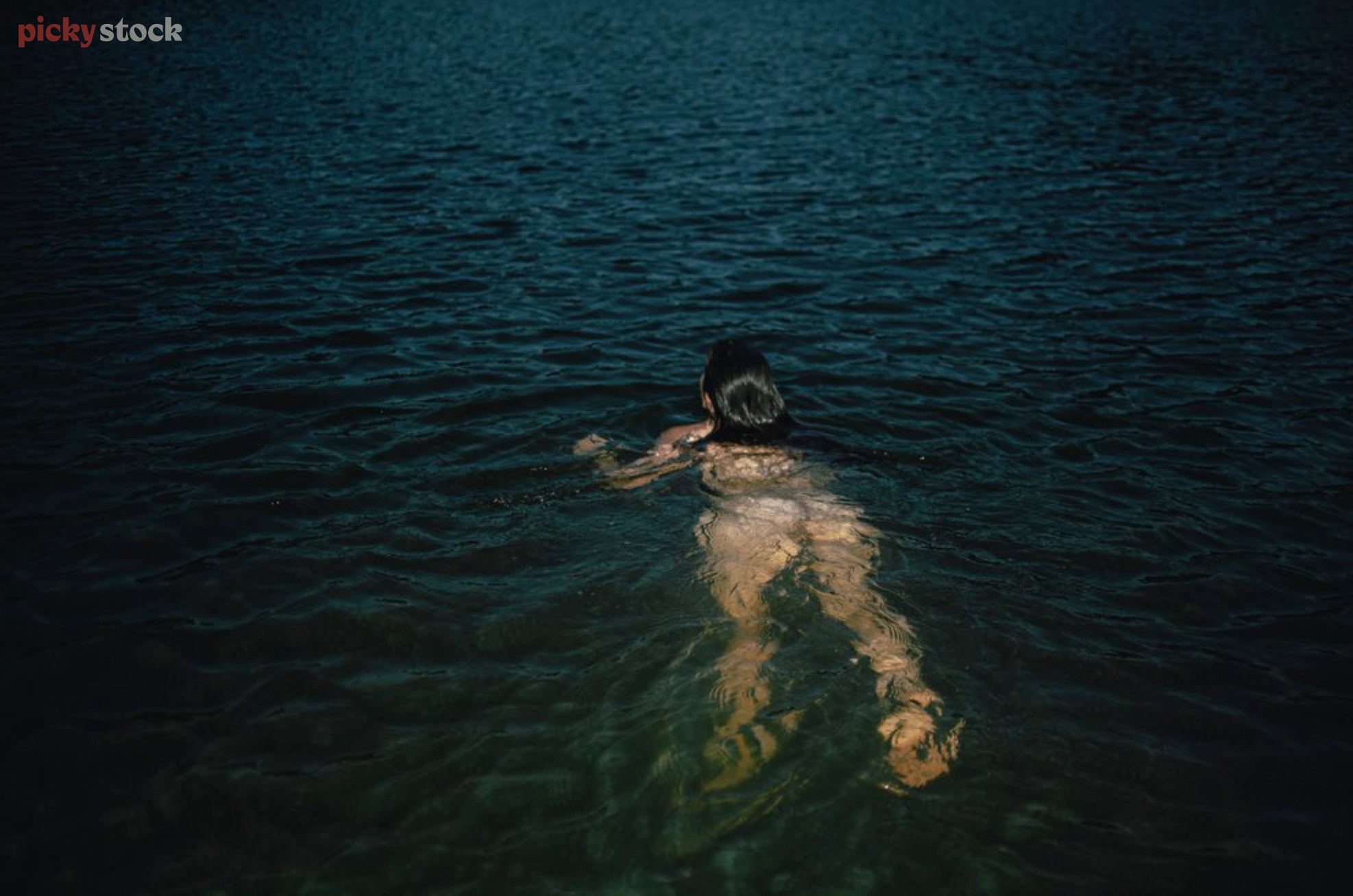 A artist shot of a lady skinny dipping in the New Zealand ocean, during a summers day. Her body is blurred by the water ripples with just her brunette hair breaking the surface. 