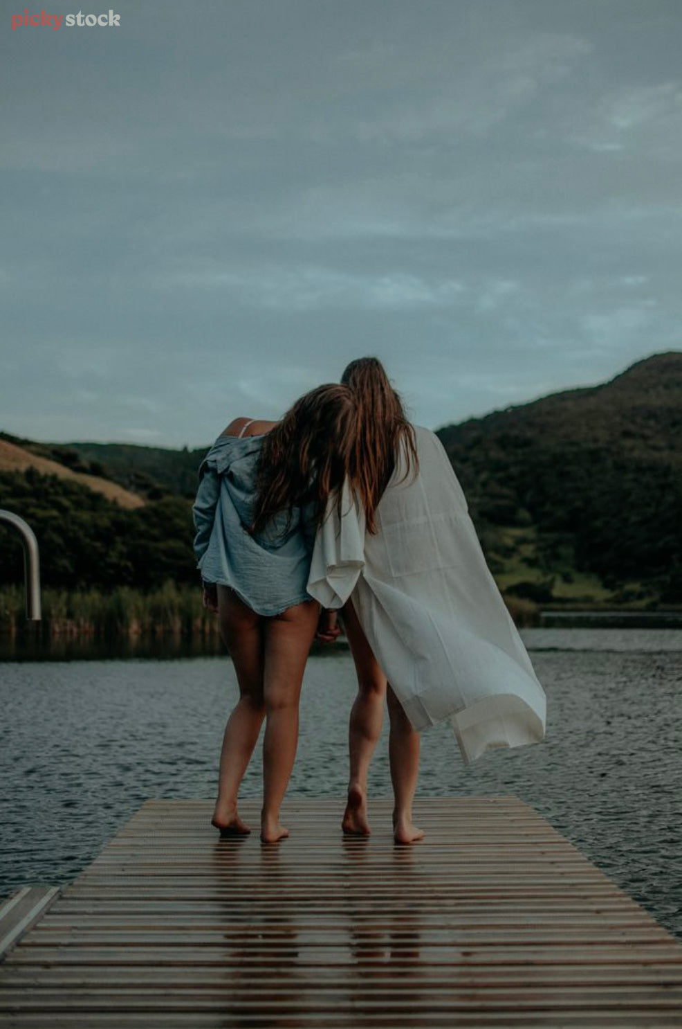 Two female friends in their mid twenties stand at the end of a jetty gazing out towards Lake Wainamu, Auckland. They both lean their heads on each other in companionship on a dark day. 