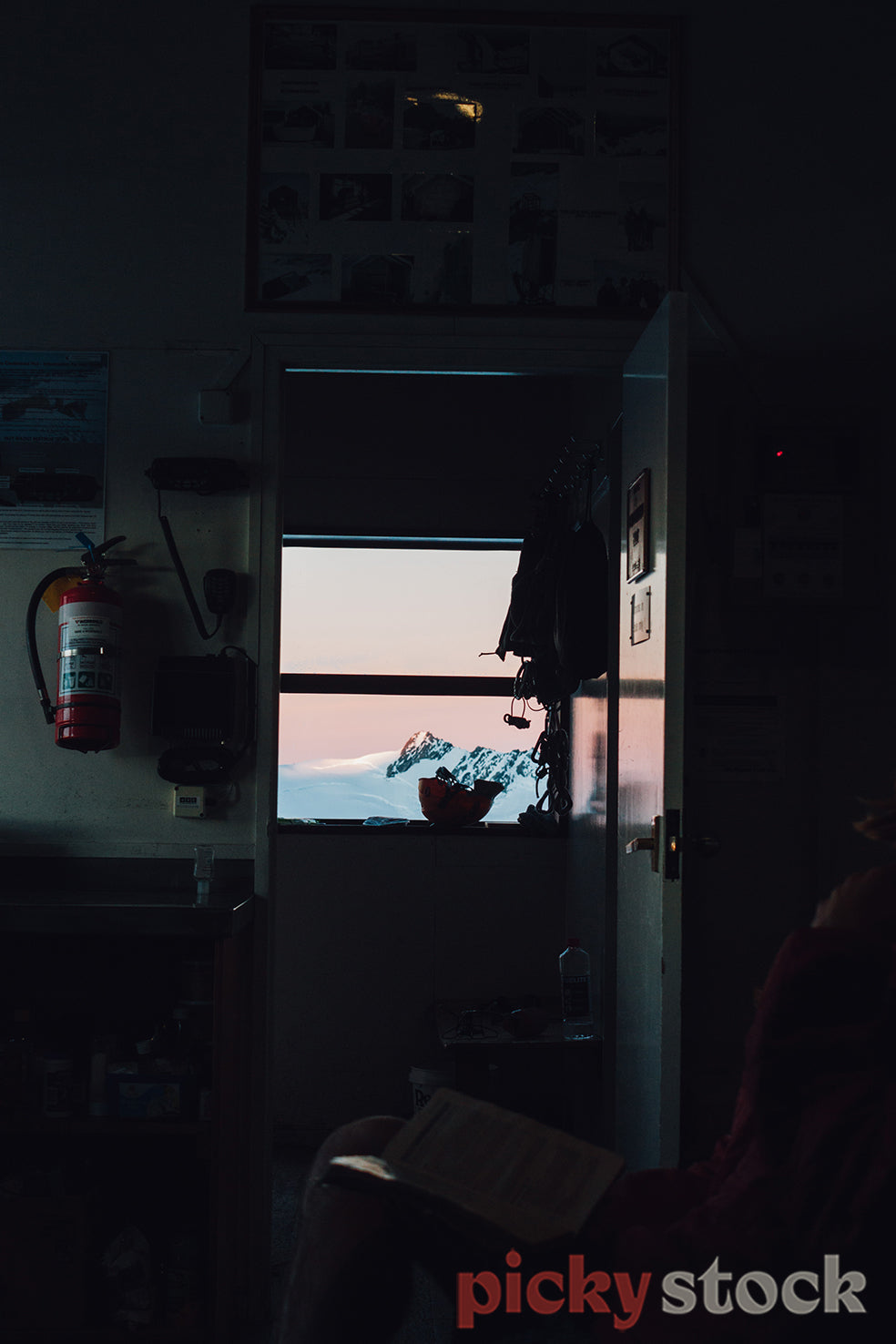 A climbing helmet is outlined on the window sill in a dark alpine hut with mountains glowing in the sunset outside the window.