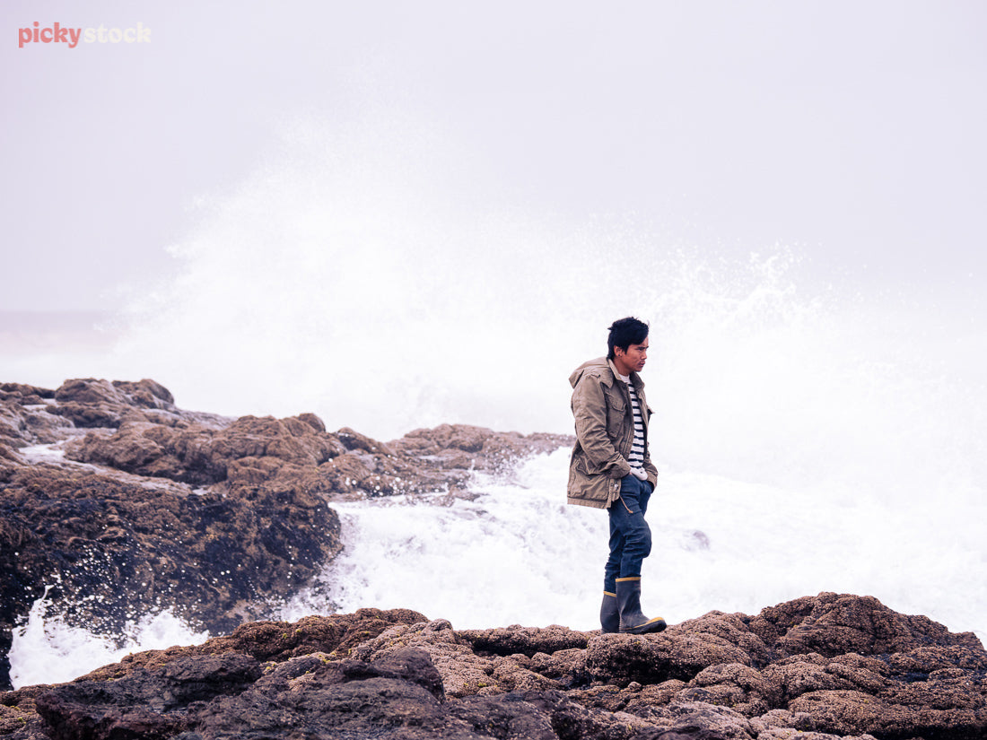 A man stands in full winter gear and gumboots on the rocks at Ninety Mile Beach, Northland, as the salty sea spray nearly splashes him. 