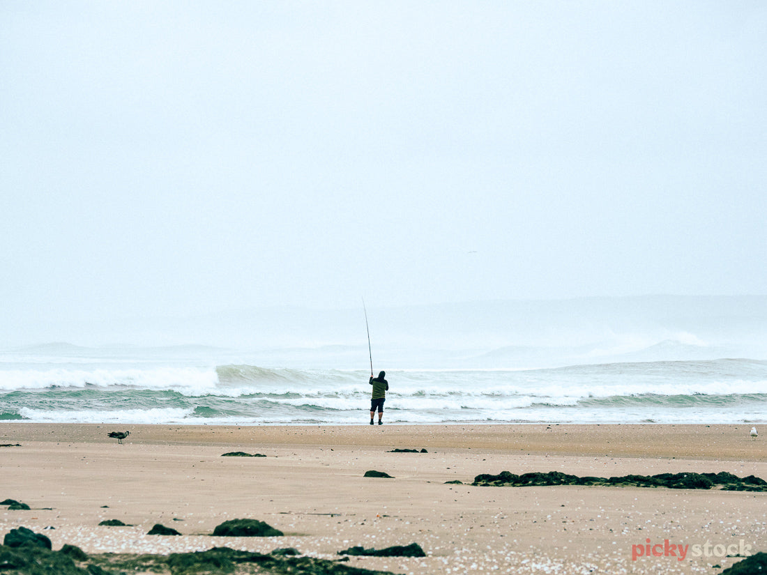 A lone fisherman stands on Ninety Mile Beach, Northland, as he casts his rod out to the messy breaking waves on a rough grey day. 
