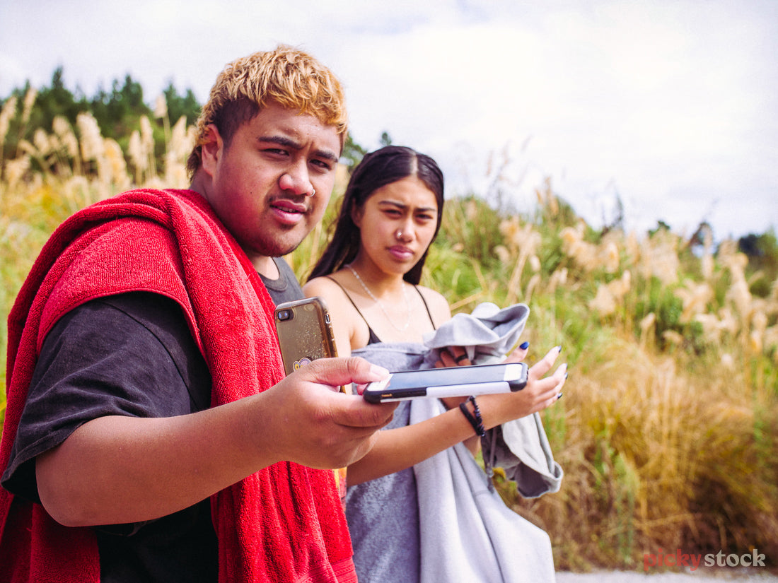 Two Maori teens look direct to camera with towels over their shoulders, ready to swim in Taupo, New Zealand. One holds out their phone to camera, as if asking someone to hold it. 