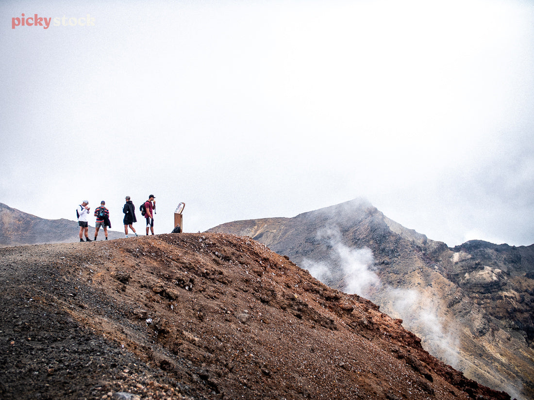 Line up of people walking along the ridgeline of Tongariro, during the Tongaririo crossing. Geothermal activity can be seen in the background of the photo, with fog rising. 