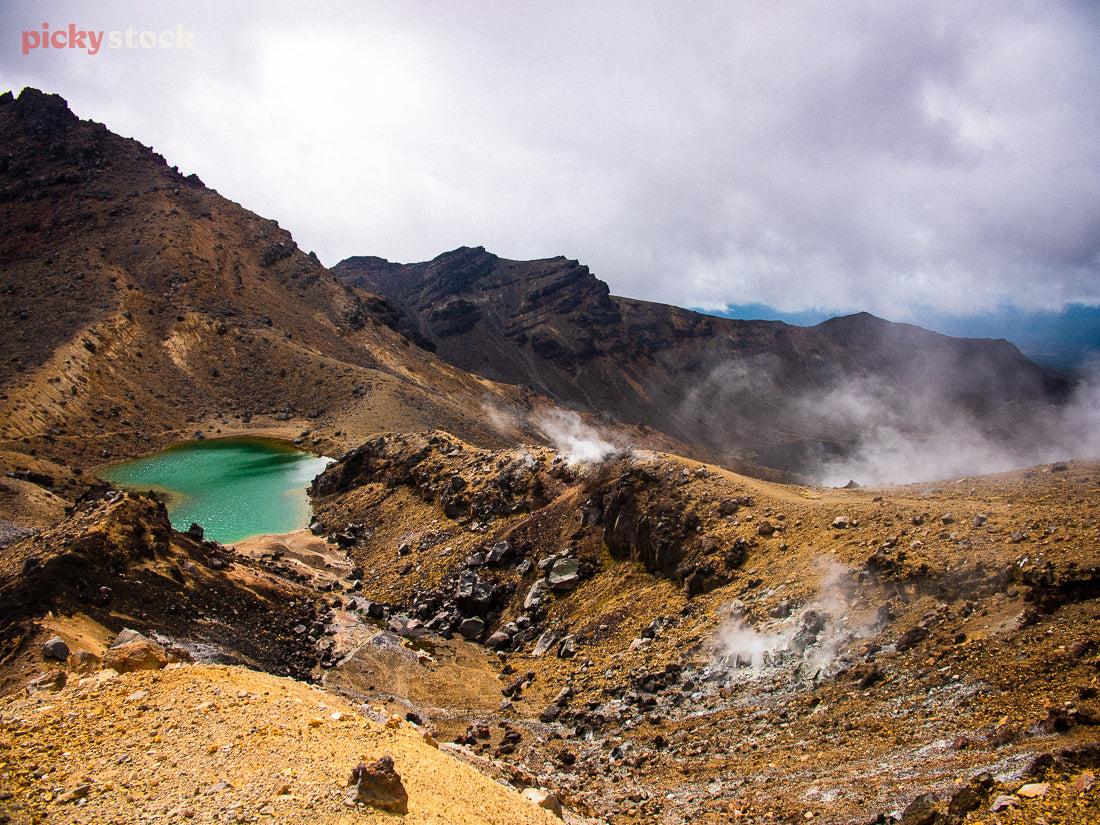 High angle image of Tongariro National Park, looking down from the ridge to the Emerald Lakes amongst the scoria. The earthy colours striking against the grey sky,  with geothermal steam rising. 