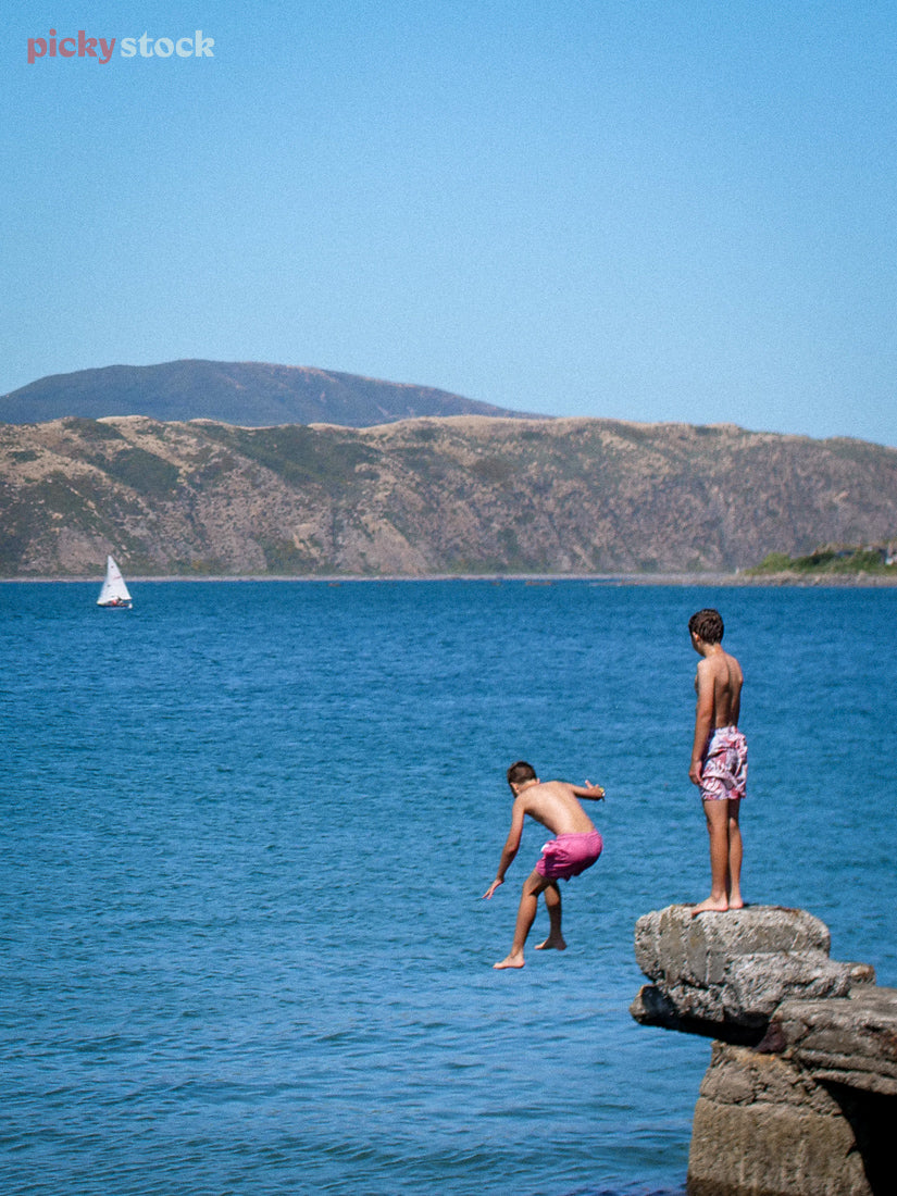 Two boys jump off a rock into the harbour in Wellington on a blue day. They both wear salmon-coloured shorts, with one boy mid-bomb and the other watching his friend jump.  