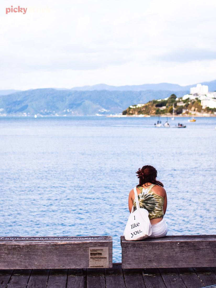 Girl sits on wooden barrier looking out at Wellington harbour on a muted blue day. Canvas tote bag over her shoulder reads 'i like you'. 