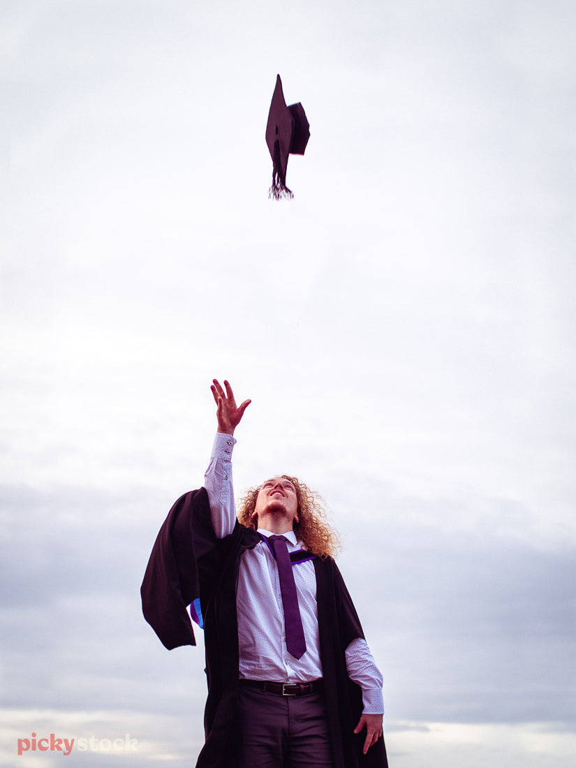 Low angle image of young adult man throwing up his graduation cap into the air. His had is ready to catch it. He wears his graduation gown over a shirt and tie. 