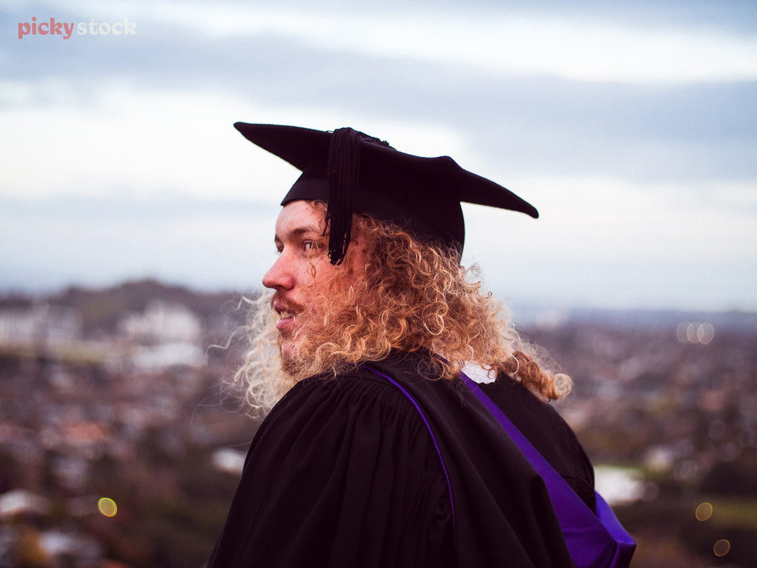 Wide angle image of a young adult man turning over his shoulder to gaze to the left. He wears his graduation cap and gown. The backround shows he is up on a hill overlooking Auckland, with the scenery out of focus behind him. 