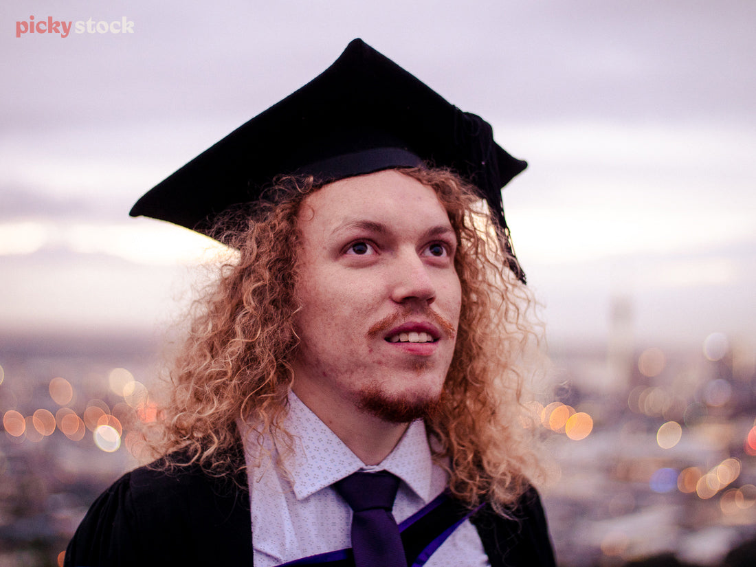 A young adult man gazes upwawrds, wearing his graduation cap and gown. The mottled early-evening lights behind him show an out-of-focus viewpoint of Auckland. 