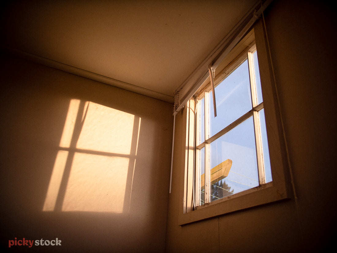 Golden morning light hits an inside wall of a house, as the strikes through the window. The day looks to be blue outside with the blinds up on the window. 