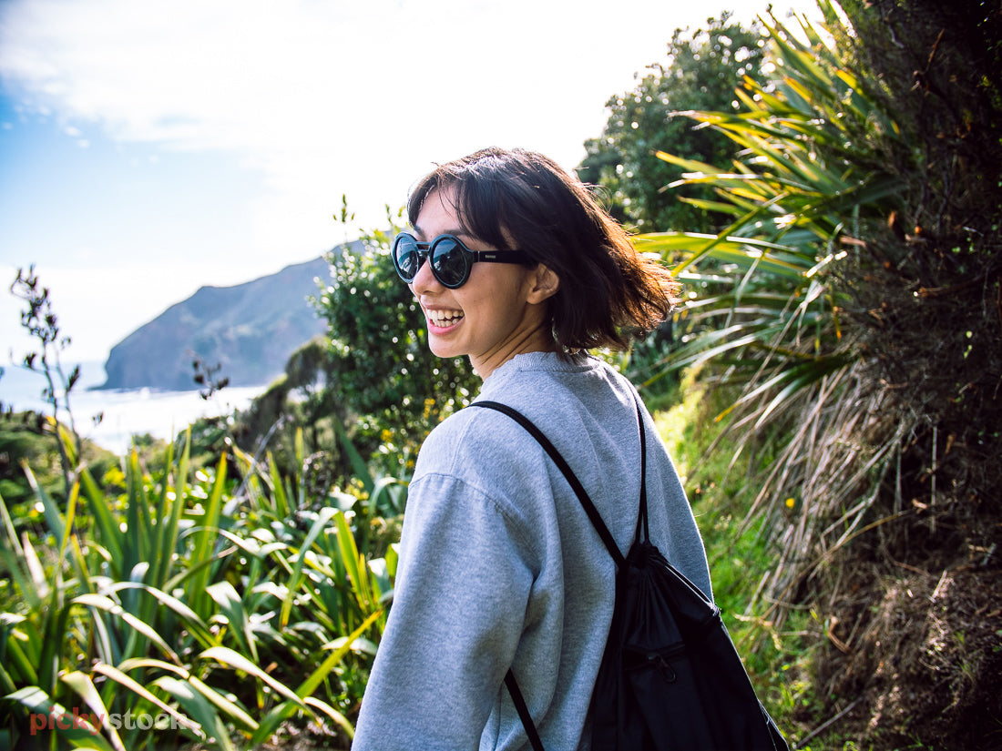 A young spritely lady walks away from camera on a coastal bush track. She has a loose backpack on and looks over her shoulder back towawrds camera. Sunglasses and big smile on her face as she goes. 