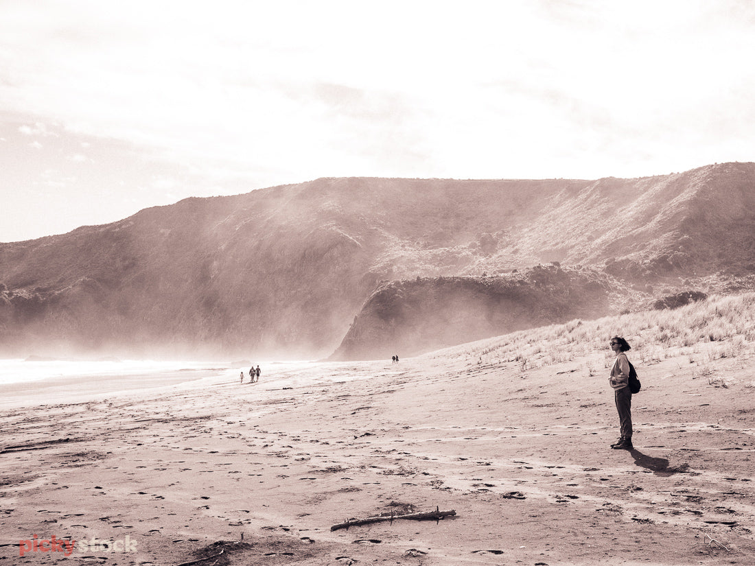 Sepia-coloured wide shot image of lady looking out from the sand to Bethells Beach, West Auckland. The sand lightly drifts upwards over the headland in the distance. 