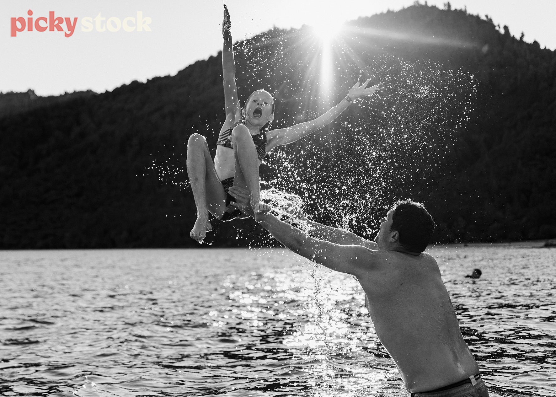 Father, man throws daughter girl wearing bikini togs swimwear out of the water. Girl has a big shocked expression on her face. Water is flying in the sky, with sunlight directly hitting her and the water in the middle of frame. They are playing in water in Blue Lake Rotorua, large mountain behind them. 