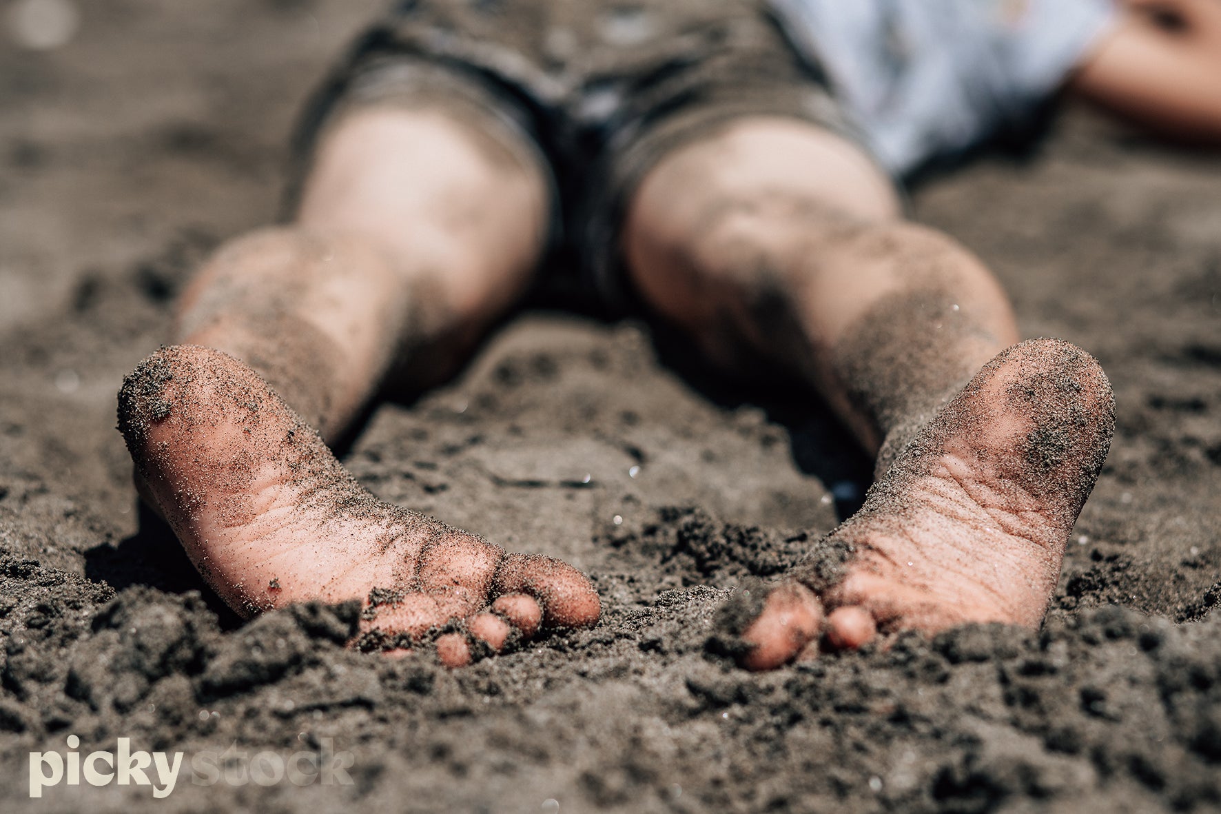 Small kid lying in a black sand beach. Close up of sandy wet small feet. Legs and body out of focus in middle of frame