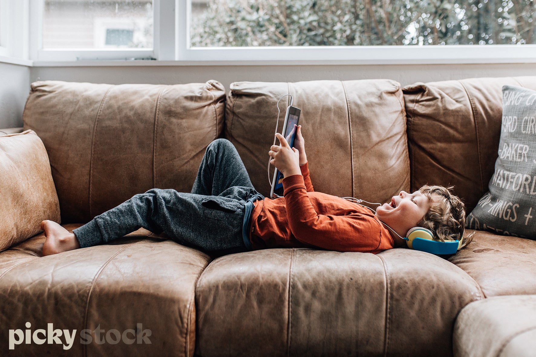 Small boy lying on his back looking at tablet / ipad wearing blue and yellow headphones. Lying on a brown leather couch. Window to the outside at the back of the image. Kid is wearing a orange long sleeve top and blue jeans with bear feet.