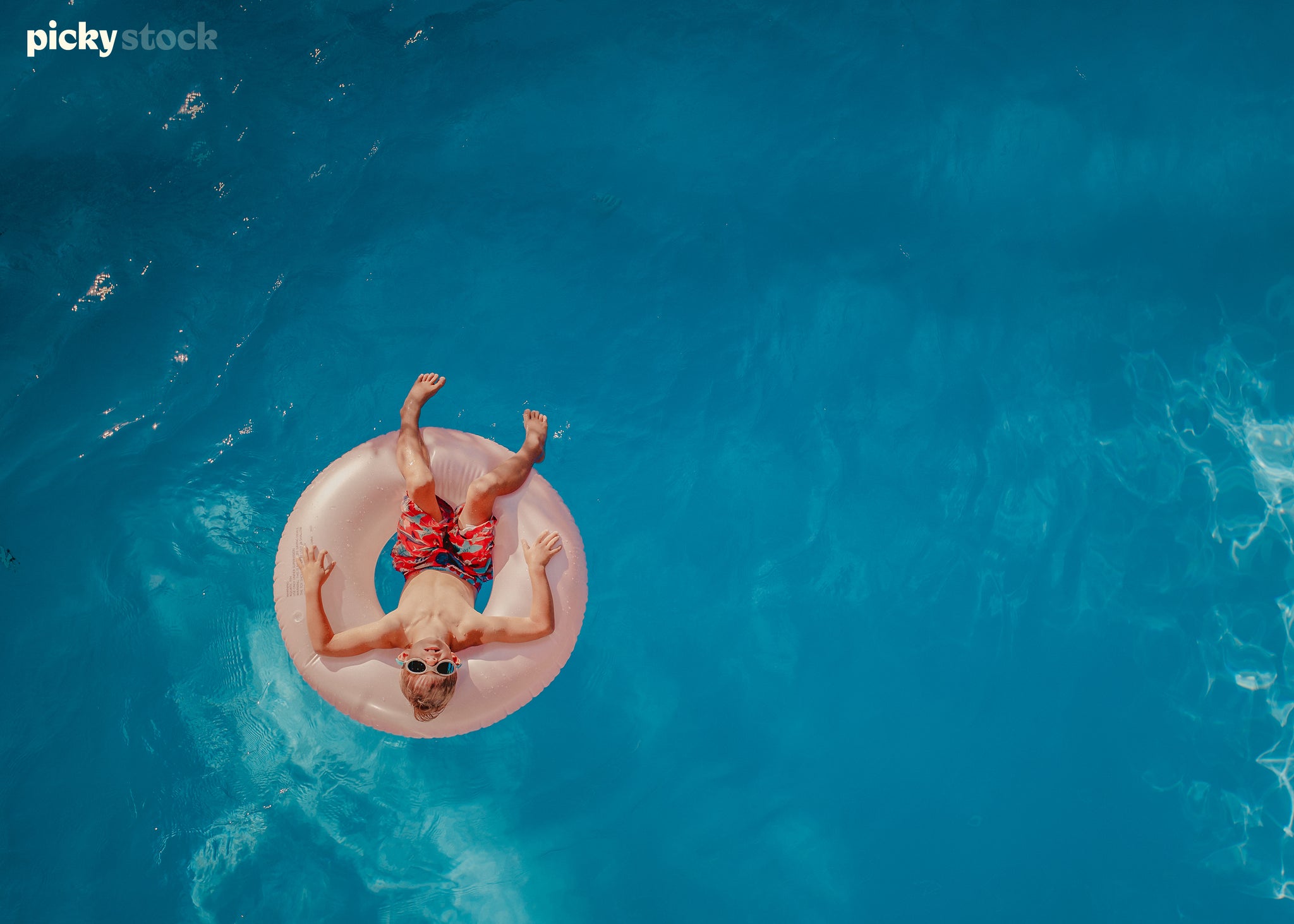 Overhead shot of a small boy with blonde hair in a swimming pool on a soft pink inflatable ring. Boy wearing red board shorts swimming togs trunks. Water is bright blue. Boy is looking up to camera wearing sunglasses