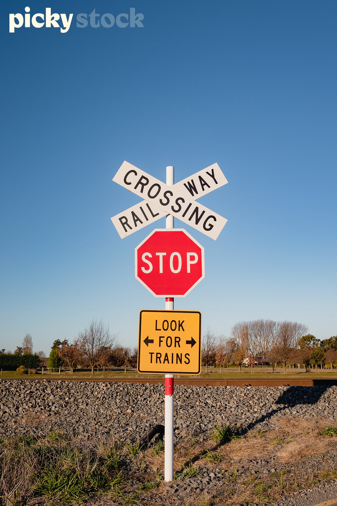 Railway crossing sign, on side of a road. Sky is blue. Stop sign, and 'Look for Train' sign in yellow also attached to crossing pole. Brown rail track in background, with gravel road around.