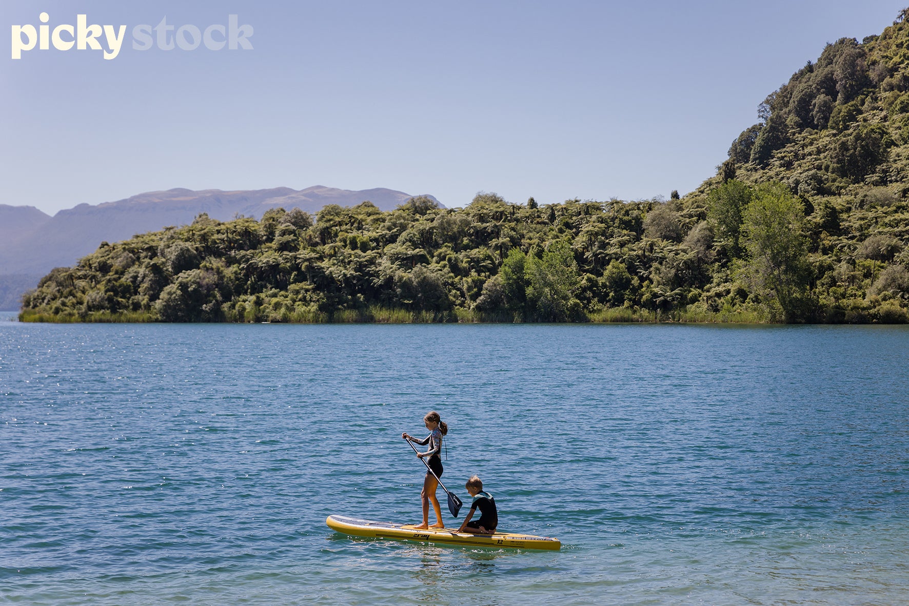 Two small kids on oner yellow stand up paddle-board. Small girl with pony tail in rash shirt is paddling. Small boy behind her. Lake is blue. Lush green cliffs surrounding the water. Blue sky, summers day.