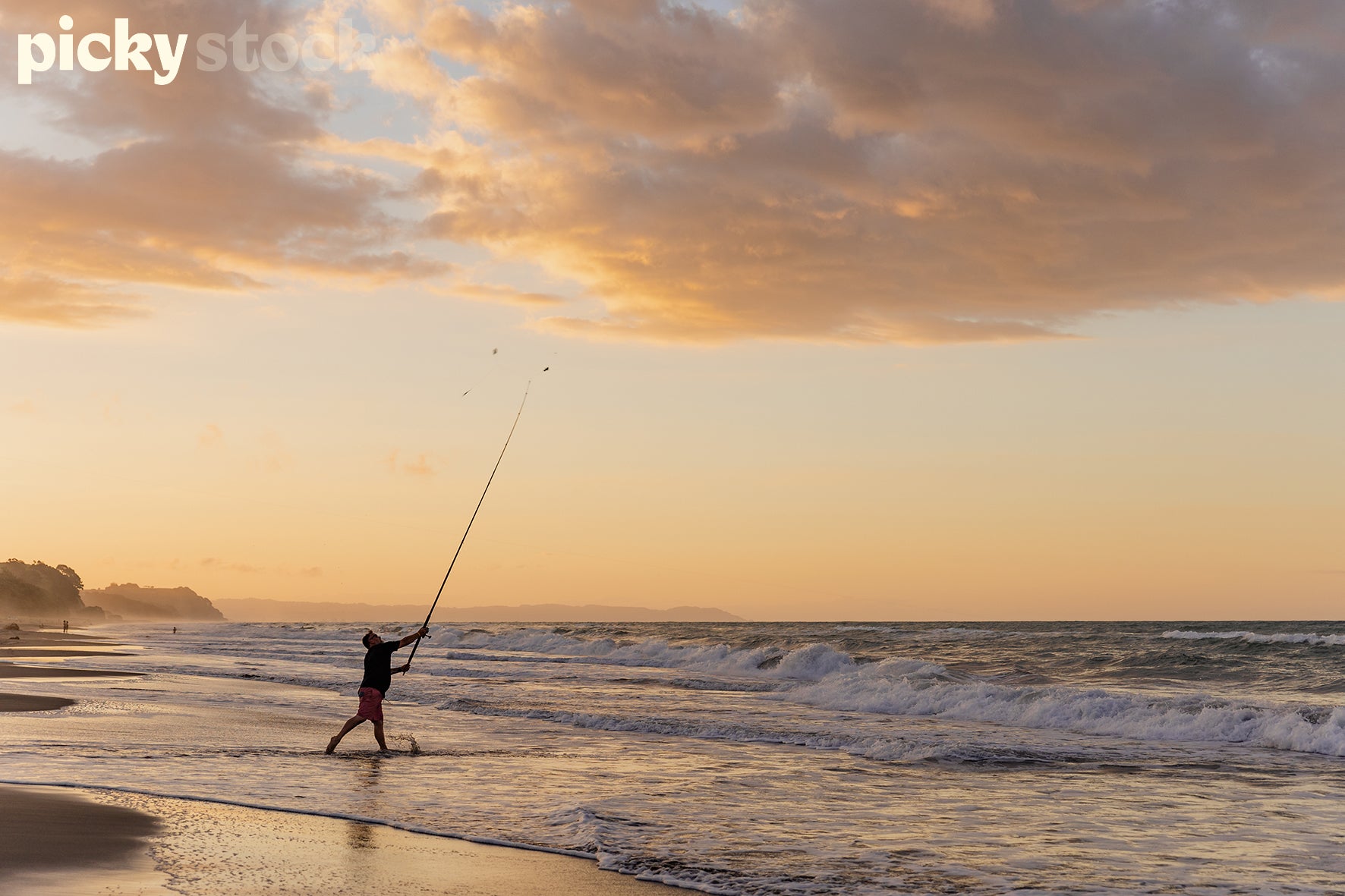 Man fishing from the beach casting his line at Opotiki Beach. Man is wearing red board shorts togs with a black long sleeve top. Fishing at sunset, with a few rolling waves breaking on the shore. Man is standing in water ankle length.