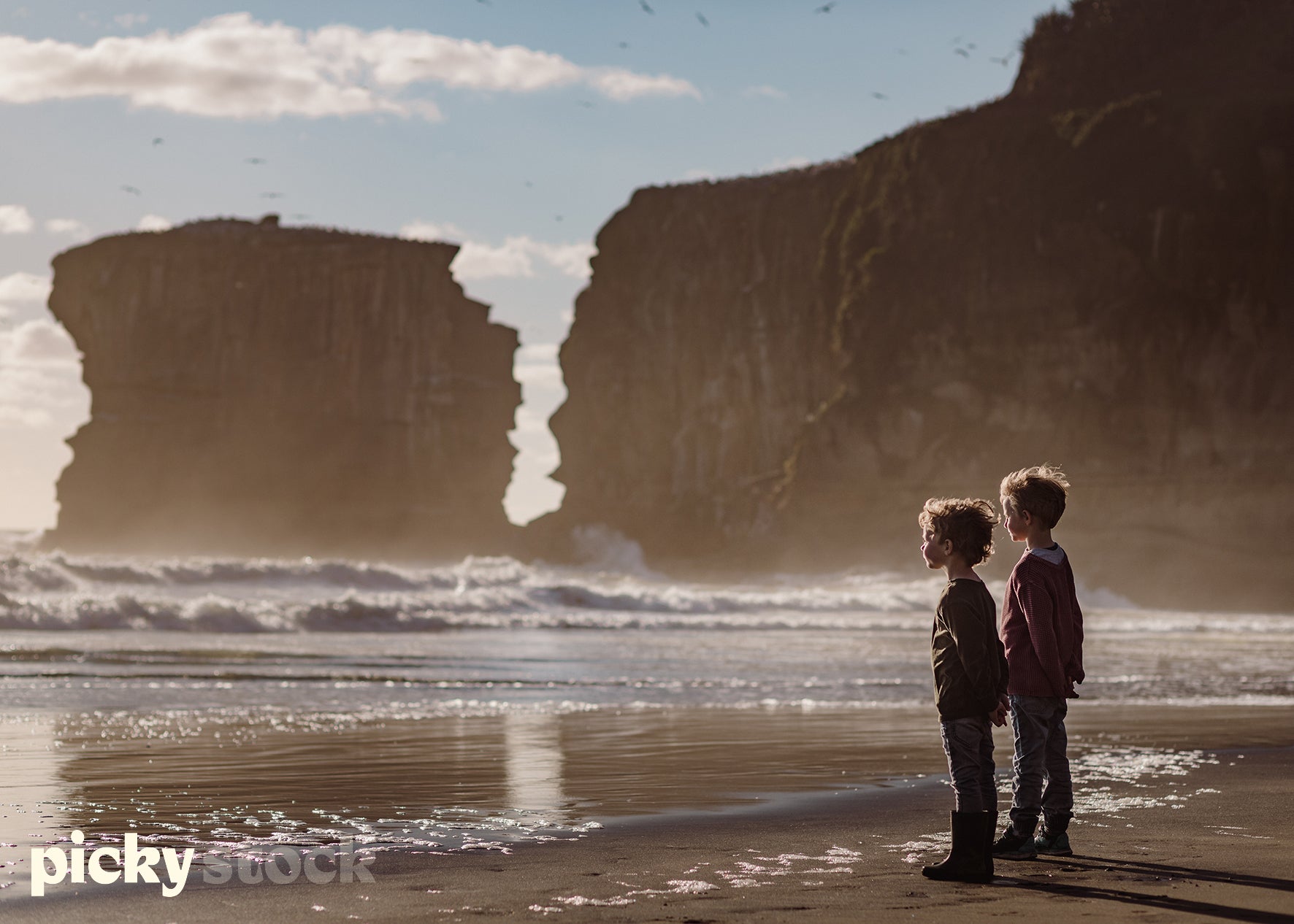 Two small young boys standing towards the ocean at sunset, watching the ocean. Reflection of each boy on the sand. Sand is back. Dramatic cliffs and rock face in the background of image. Ocean is rough with white caps and waves visible. West Coast NZ Beach
