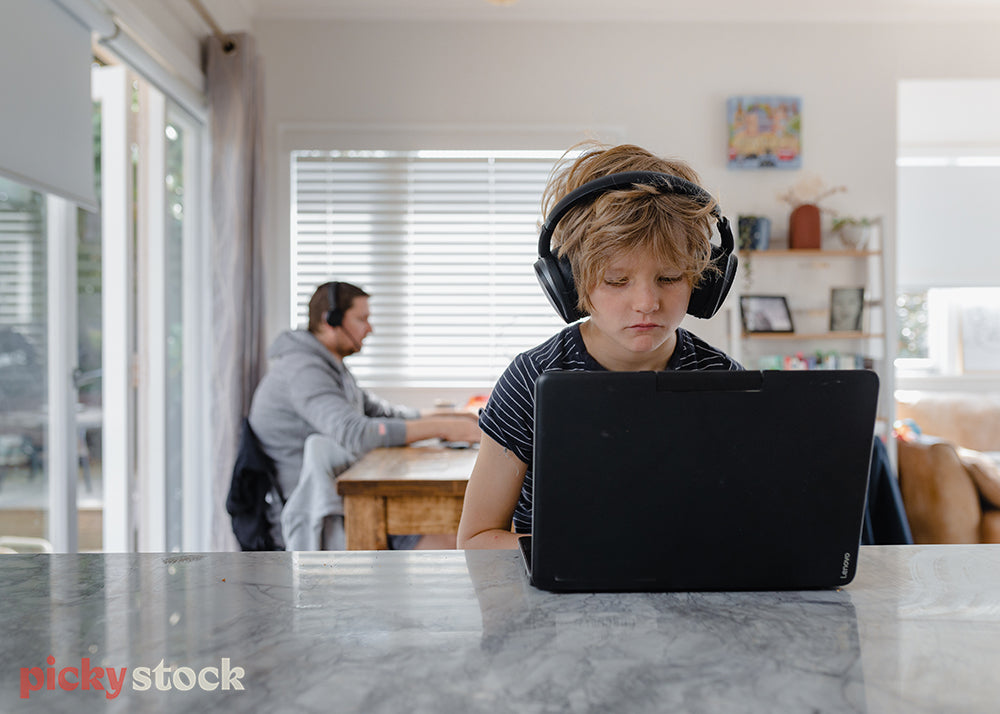 Small boy child working at marble kitchen bench with black headphones on. Laptop is black open facing him. Father is in the background to the left of him, wearing. a grey hoodie, also wearing a headset sitting at the kitchen table. 