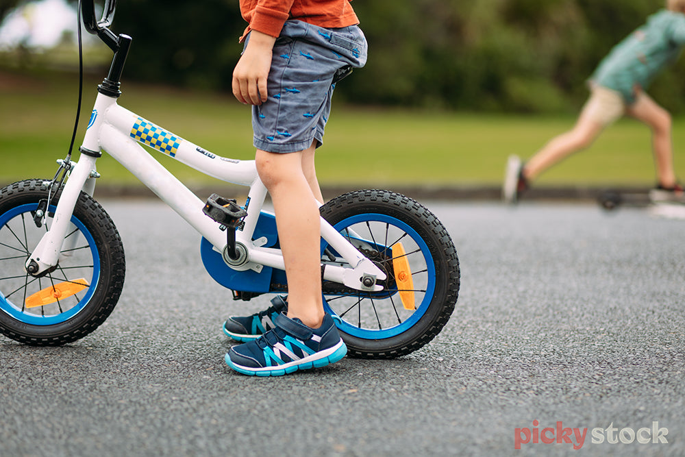 Faceless image of boys riding a bike and scooter. Boy is wearing blue shoes, camera cropped tight at boys hips. Bike is white with small checker details. 