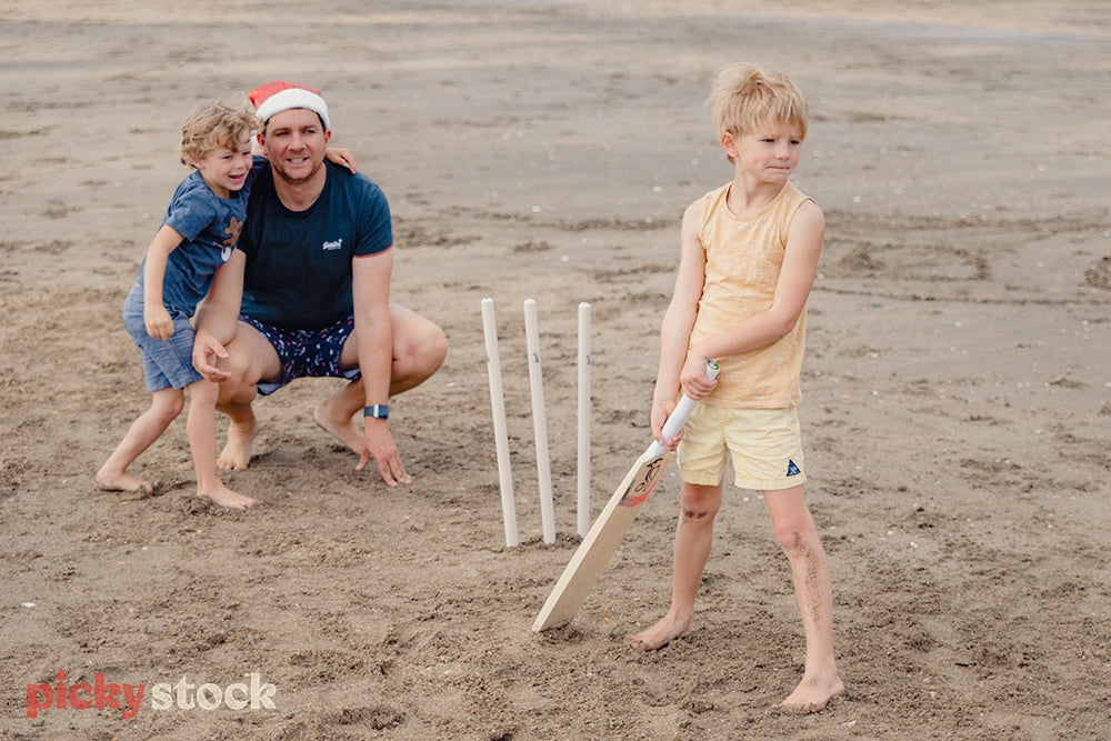 Father and two young sons playing cricket on Christmas Day. Blonde boy is holding the cricket bat wearing yellow top and shorts. Dad wearing santa hat and blue top and board shorts with small son watching with him behind the wickets.