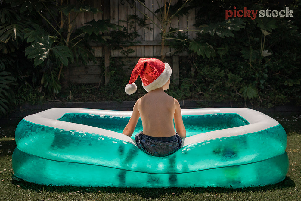 Faceless image of young boy sat on a florescent aqua green paddling pool with Santa hat on. Boy is in the middle of the frame, sitting in the middle of the pool. Pool is pushed down holding his weight. Greenery and fence behind. 