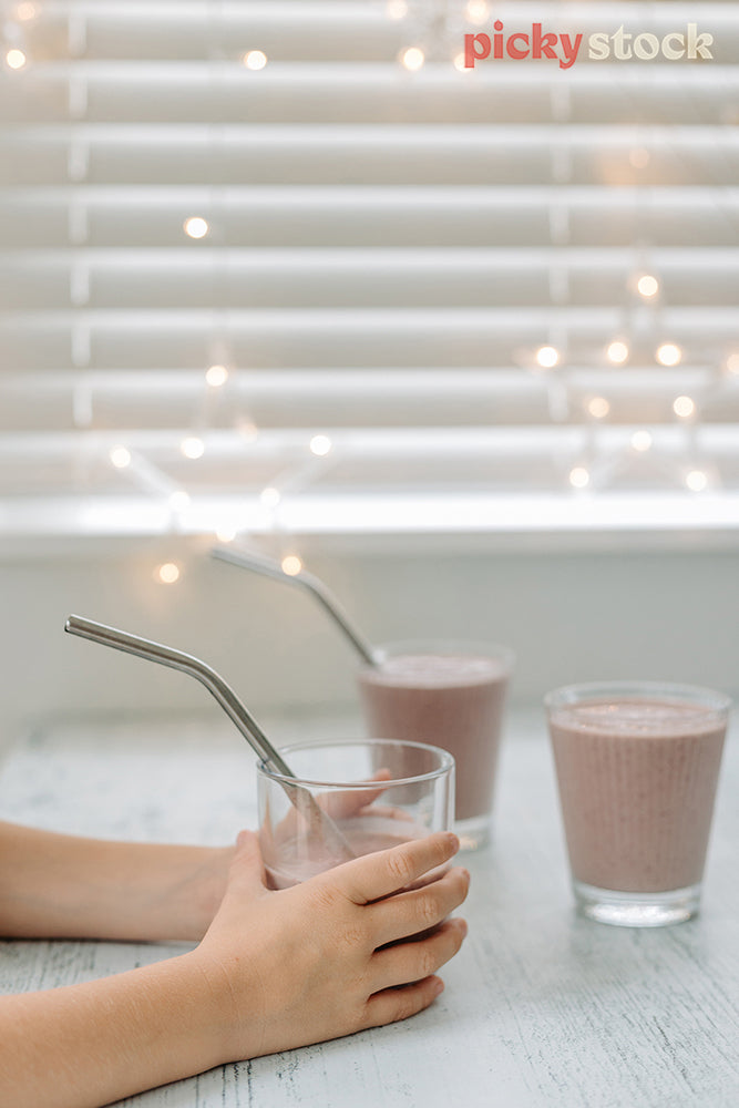 Small hands holding a smoothie with a metal drinking straw. White blinds and fairy lights in the background. three smoothie glass cups on the bench, filled with strawberry smoothie. A metal straw in each. 