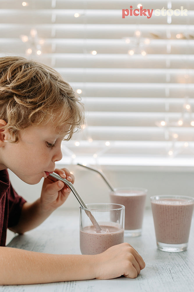 Young blonde boy left of frame, holding metal straw drinking from a glass. He is looking down and sucking the drink. The smoothie is strawberry and in a clear glass.  White blinds and fairy lights in the background. three smoothie glass cups on the bench, filled with strawberry smoothie. A metal straw in each
