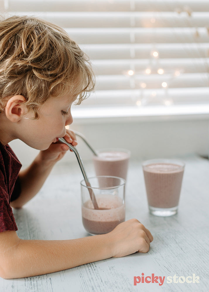 Overhead shot of young blonde boy left of frame, holding metal straw drinking from a glass. He is looking down and sucking the drink. The smoothie is strawberry and in a clear glass. White blinds and fairy lights in the background. three smoothie glass cups on the bench, filled with strawberry smoothie. A metal straw in each