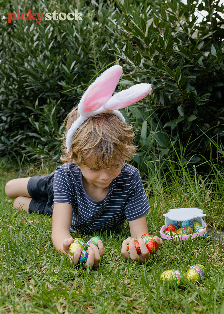 Portrait image of small boy wearing pink bunny ears looking at his hands, holding the Easter eggs he has collected so far. 