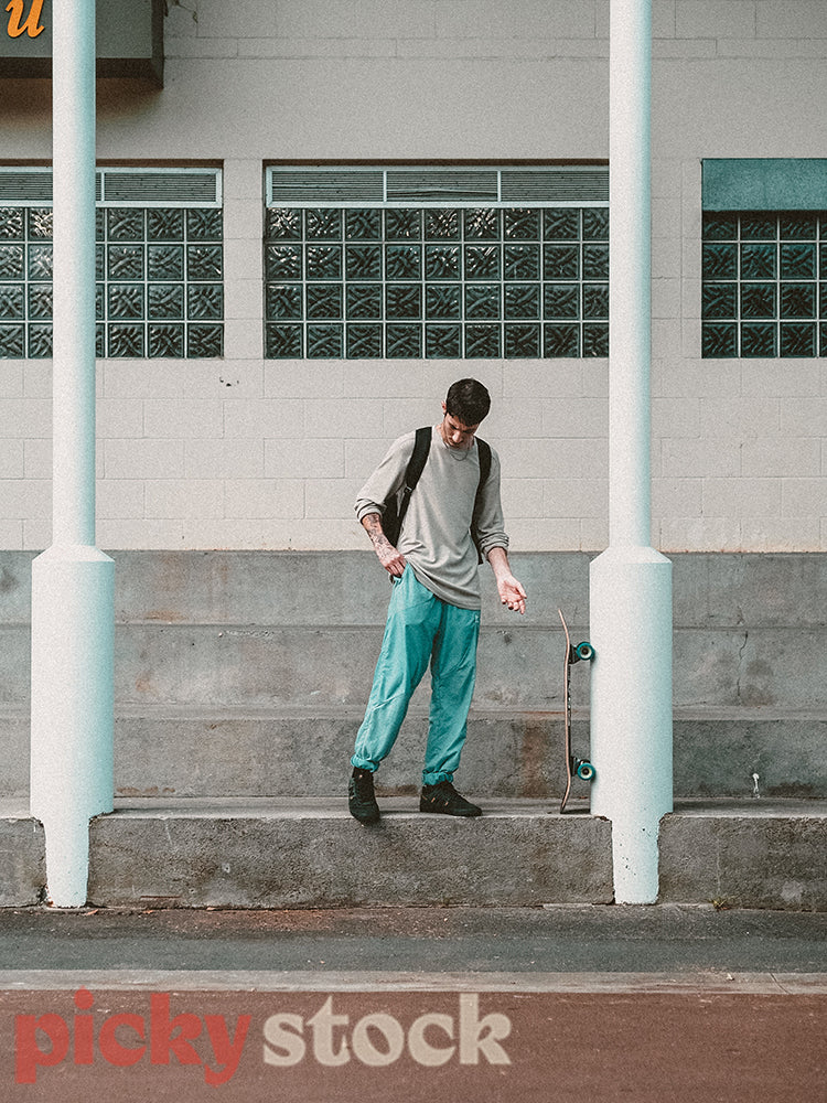 Portrait of a young adult man with his longboard, wearing a backpack and standing between two poles.
