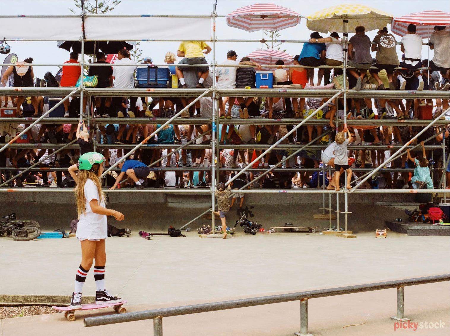 A young girl in a retro outfit and green helmet skates on sand behind a grandstand event crowd. She casually has her hair out and looks effortless while everyone else on the grandstand behind her looks to be watching something in front of them. There's retro coloured umbrellas dotting the stand and another child climbs the railing to also watch. 