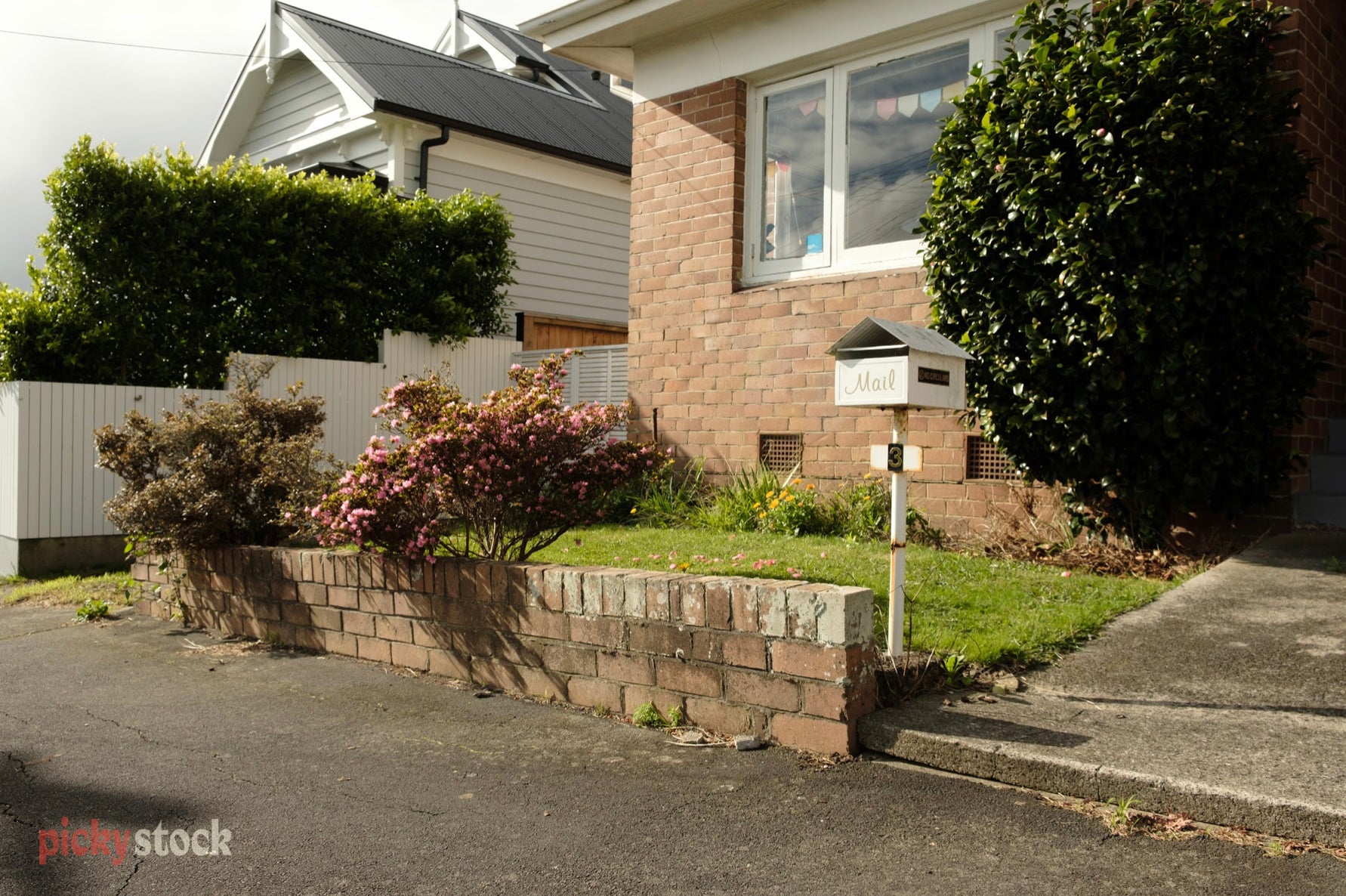 A landscape of suburban orange brick house with a white metal mailbox bearing the number three and a small brick retaining wall with a garden blooming atop it.