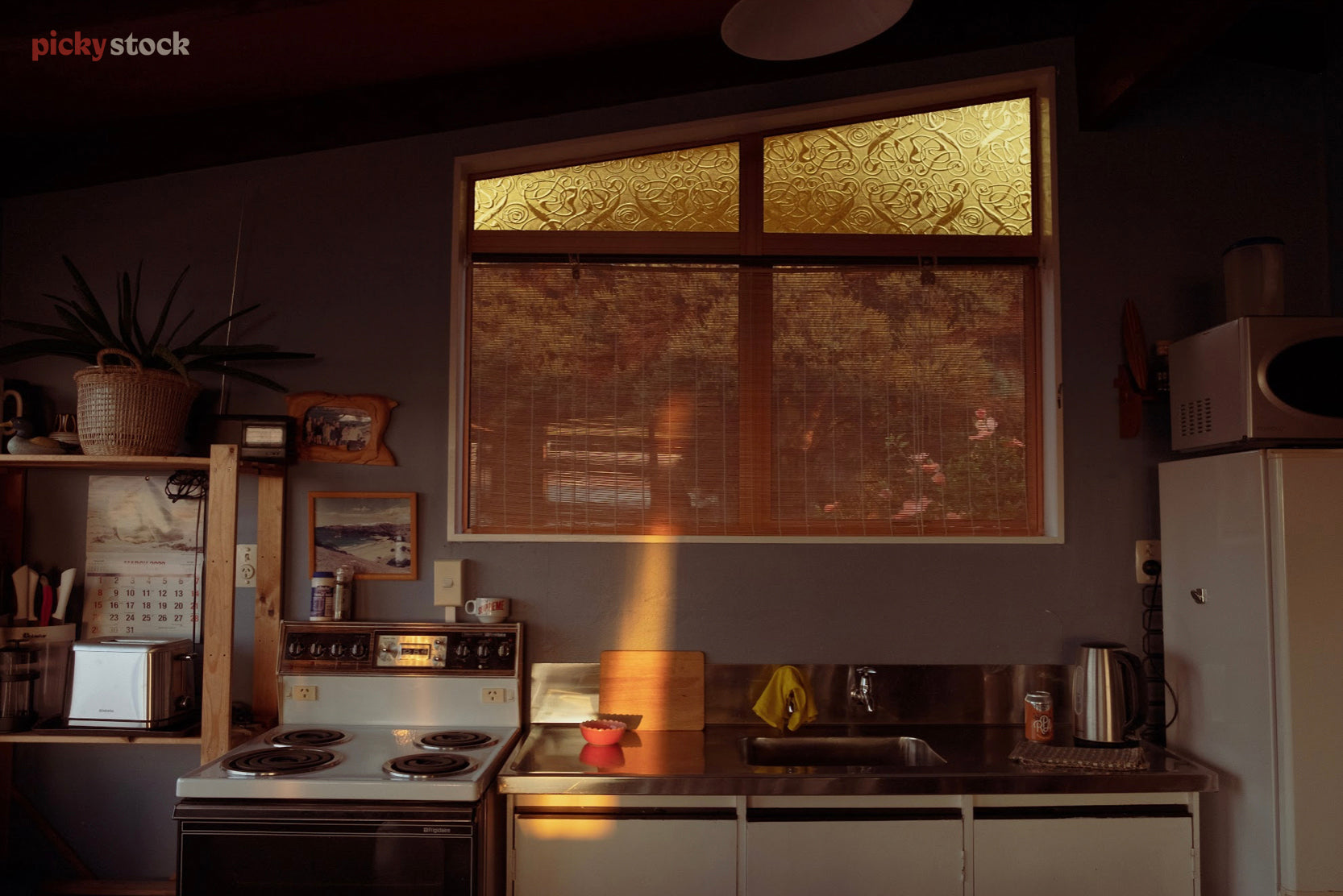 Landscape of a sun-cloaked kitchen in a mid-century style with some modern appliances spread throughout. A single slanted window casts beams of angled light over the countertop.