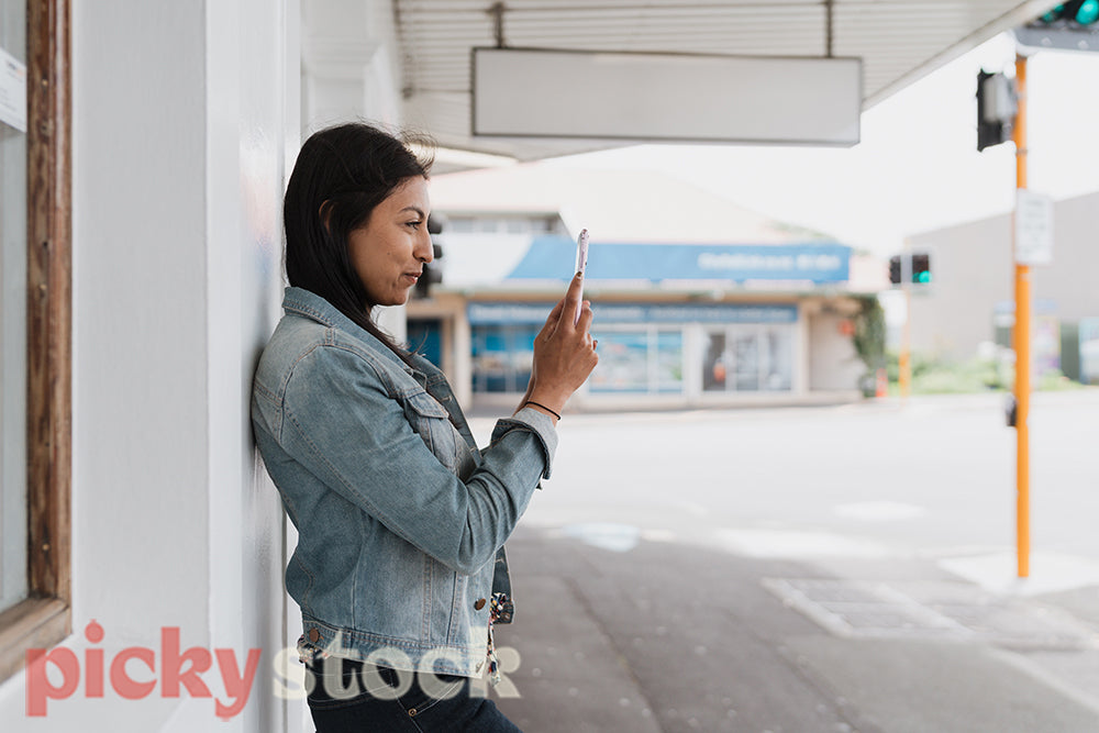 An Indian woman in jean jacket checks her mobile phone leaning against a white shopfront.