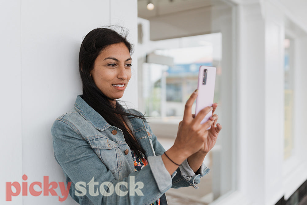 An Indian woman in jean jacket checks her mobile phone leaning against a white shopfront.