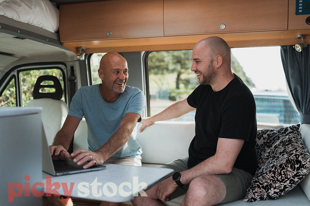 Two men use a laptop while sitting in a van with trees and beach in the background