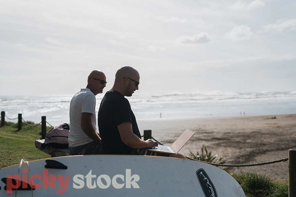 Two men sit on the beach with a surfboard next to them using a laptop while the waves break behind them.