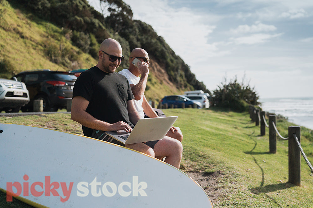 Two men sit on the beach with a surfboard next to them, one using a laptop the other taking a phone call on his mobile phone while the waves break behind them.