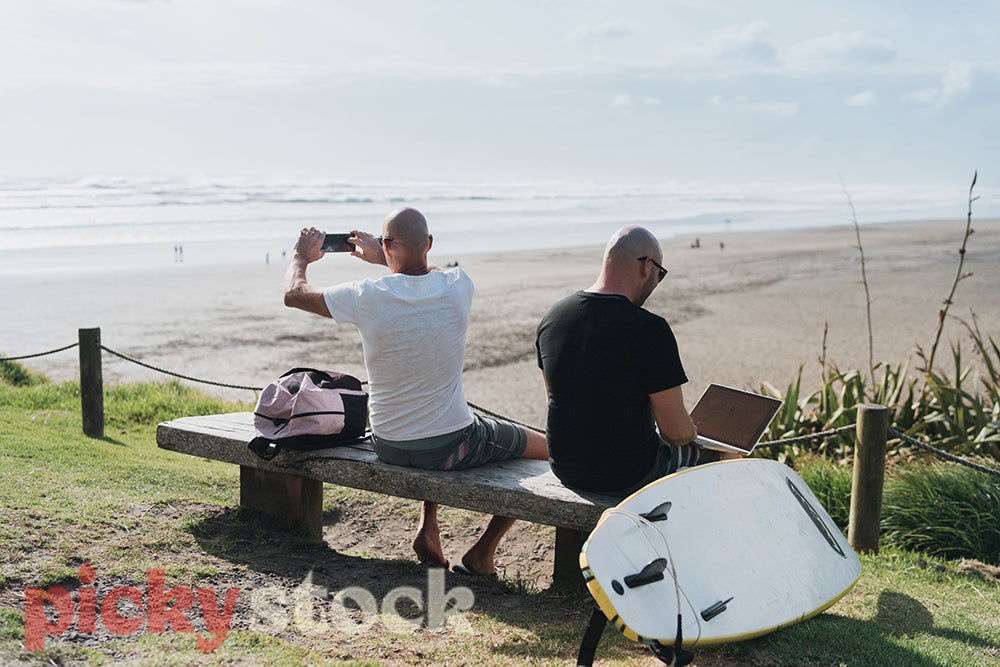 Two men sit on the beach with a surfboard next to them, one using a laptop the other taking a photo while the waves break behind them.