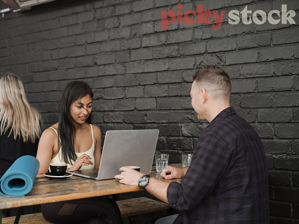 An indian woman and a man sit at a cafe with a coffee against a black brick wall. She is working on her laptop and has a foam yoga mat with her.