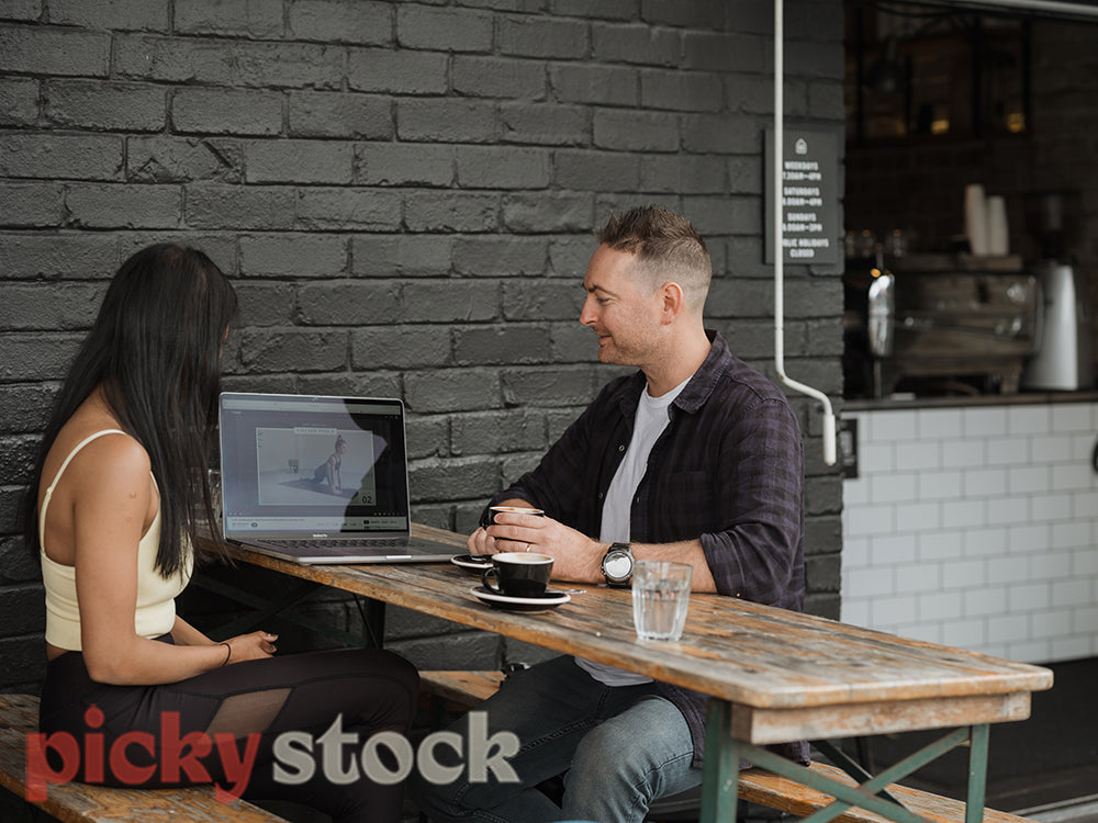 An indian woman and a man sit at a cafe with a coffee against a black brick wall. They are discussing what is on a laptop screen.