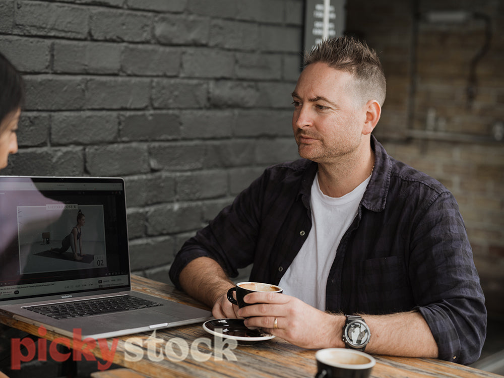 A man and another person sit at a cafe with a coffee against a black brick wall. They are discussing what is on a laptop screen.