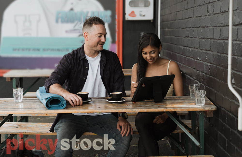 A man and another person sit at a cafe with a coffee against a black brick wall. They are discussing what is on a laptop screen.