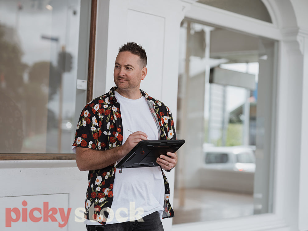 A man in a hawaiian shirt checks a tablet device on an urban city street in front of a white store front
