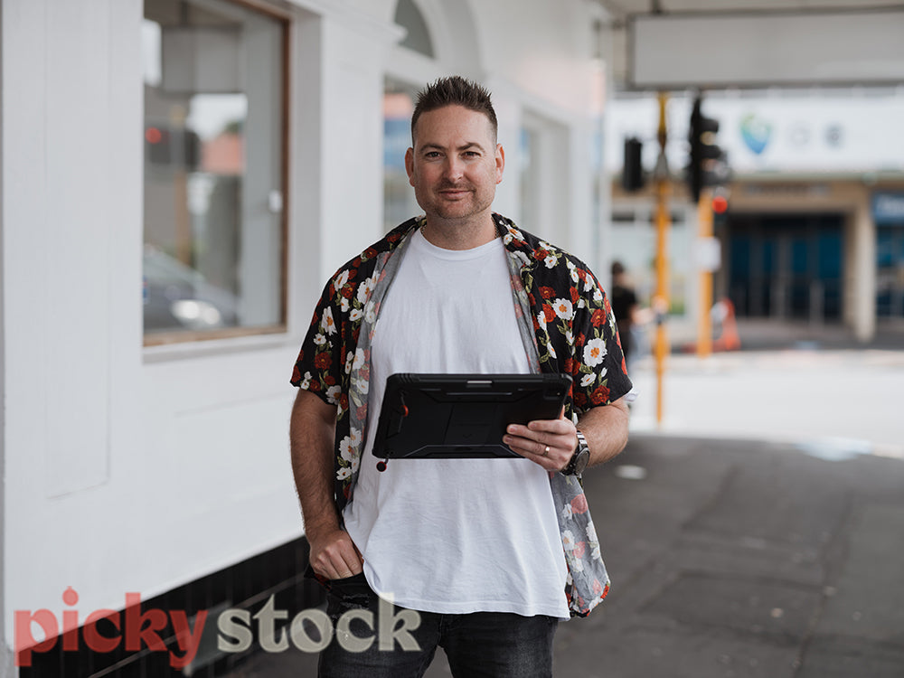 A man in a hawaiian shirt checks a tablet device on an urban city street in front of a white store front