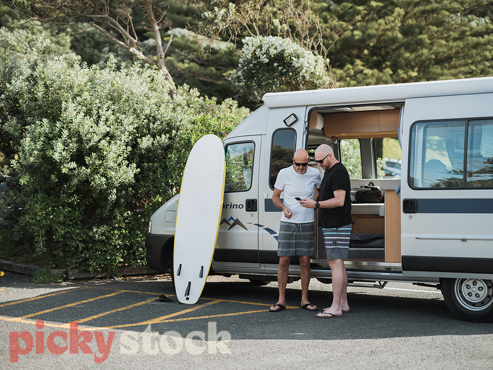 Father and son standing outside parking caravan, looking down at mobile devices. Surf board standing against passenger window.