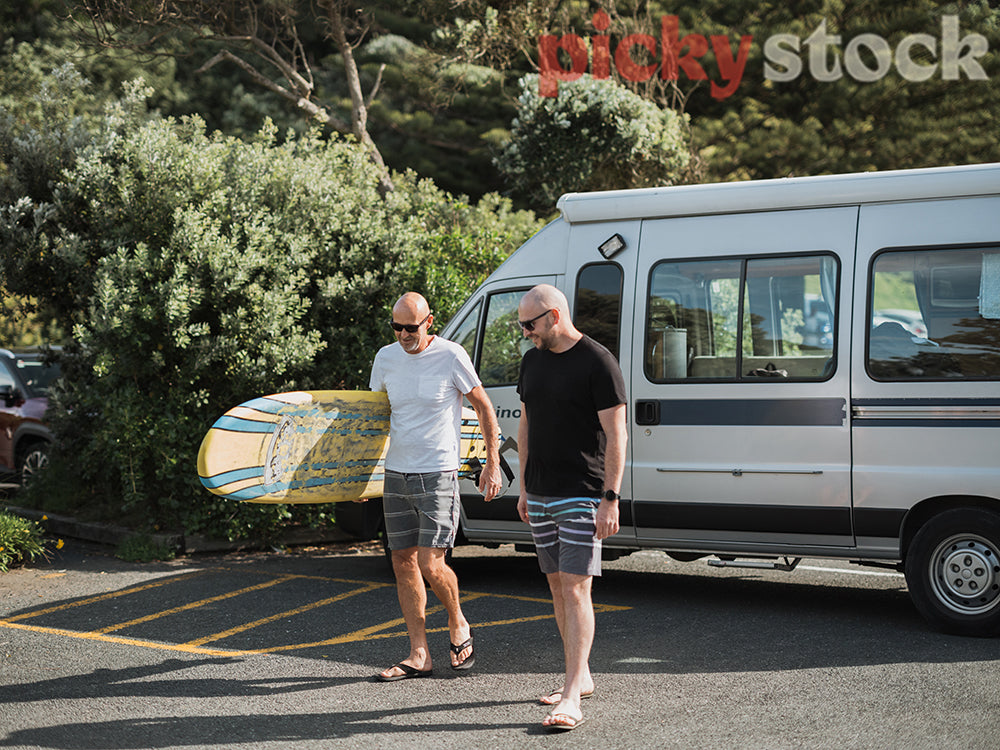 Two mean, one holding surf board, walking in beach clothes towards camera. Caravan behind them